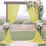 An ivory chiffon 4m x 4m stage backdrop drapes a wooden wedding arch adorned with white and peach flowers, set on grass with scattered petals and greenery in the background.