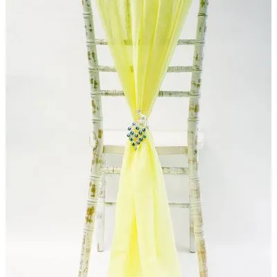 A rustic white wooden chair features the Chiffon Chiavari Vertical Drop Light Yellow—a sheer yellow sash gathered at the centre with a crystal brooch for elegant wedding chair décor, shown against a plain white background.